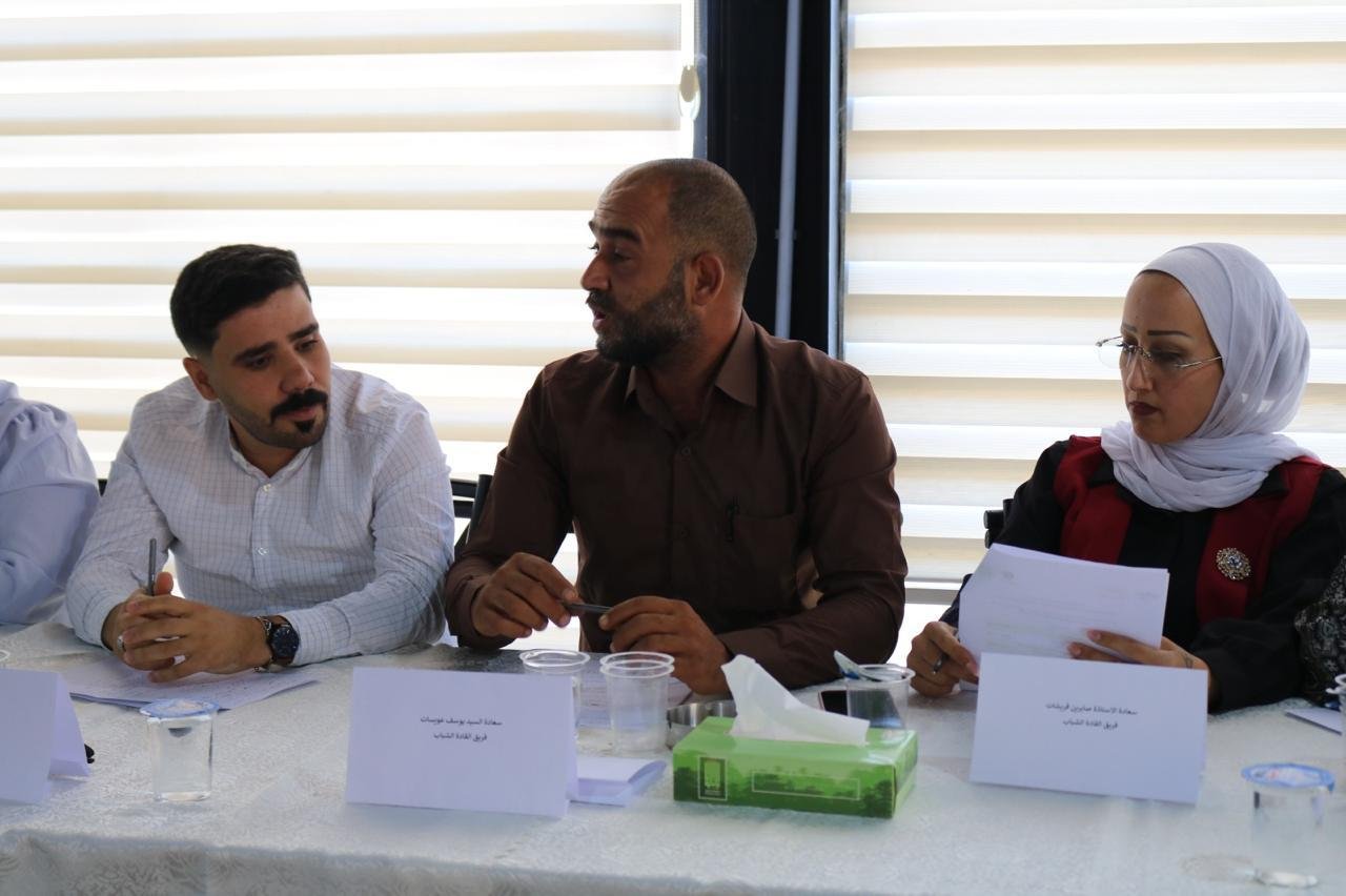 Three people sit at a conference table, two men and a woman in a hijab, engaged in discussion with papers and name cards in front of them.