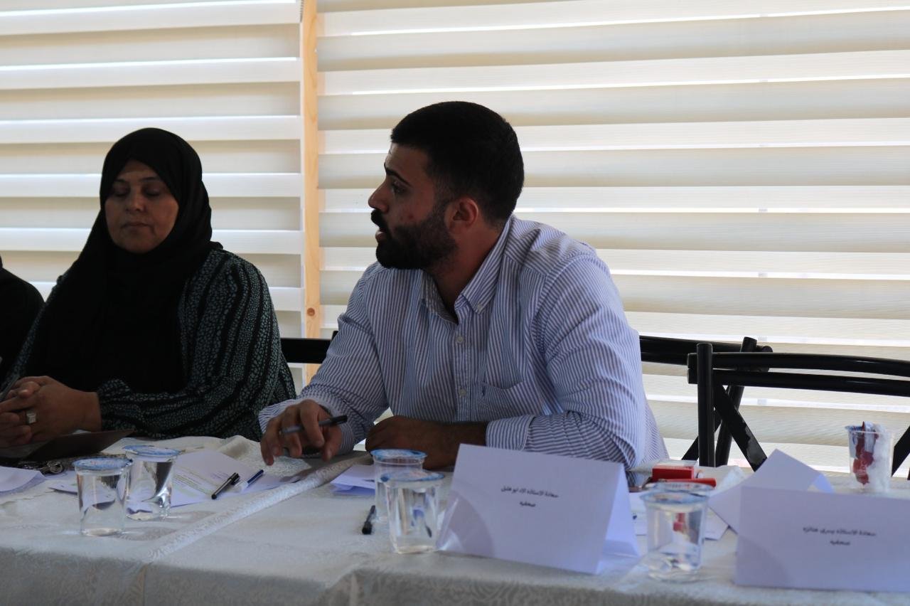 Man with a beard in a striped shirt sits at a conference table, speaking, with a woman in a hijab beside him; papers, pens, and name cards on the table.