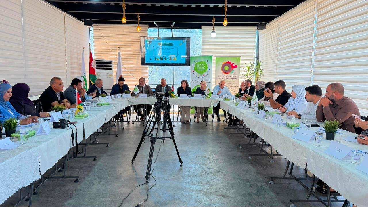 People sit around a U-shaped conference table in a bright room with banners and flags in the background, a tripod with camera in front suggests a formal meeting.