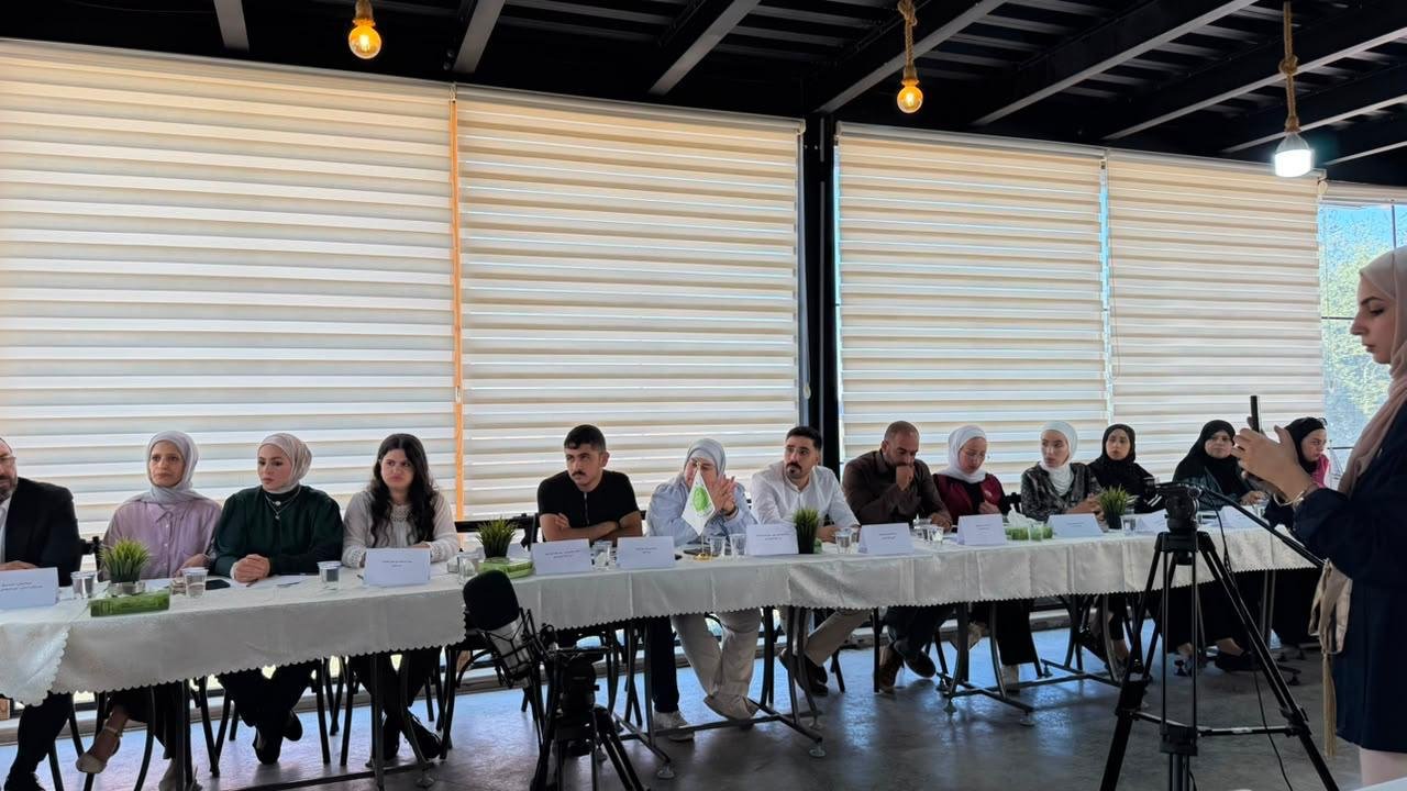 Panel of diverse people seated at a long table with name cards during a meeting, a woman in a hijab records with her phone at right side.