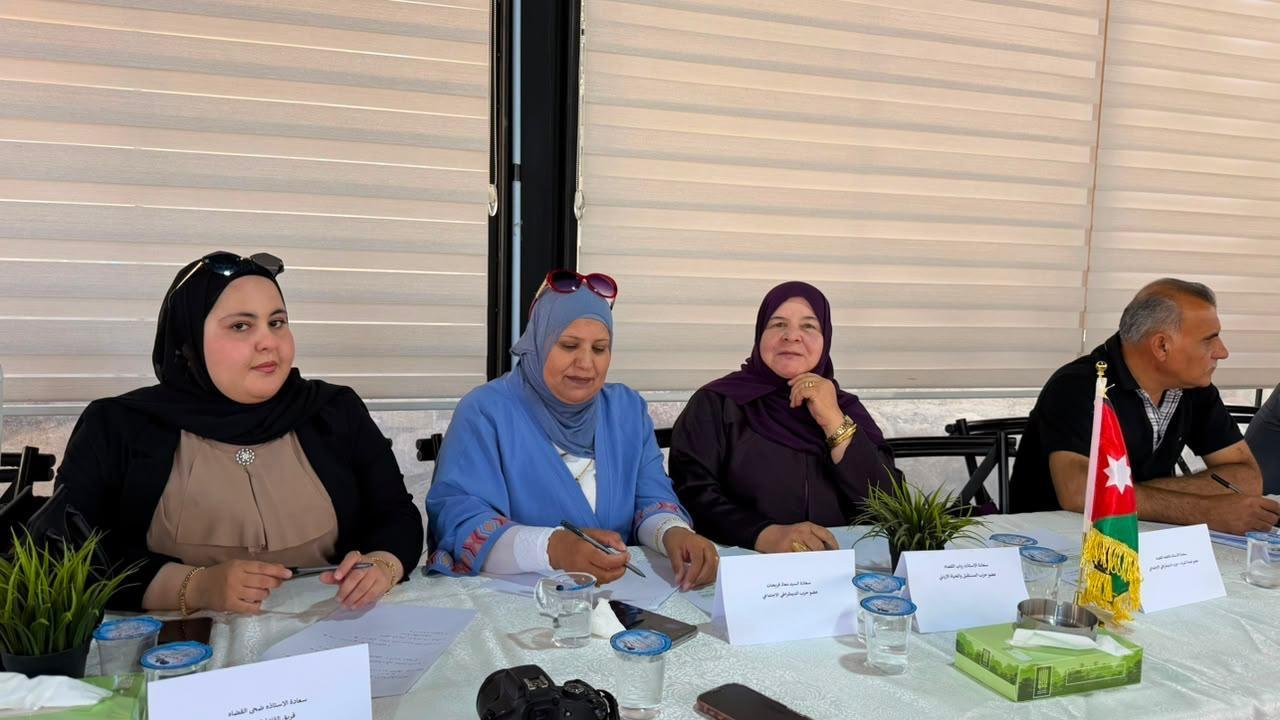 Four adults seated at a meeting table with papers, pens, and water glasses, a Jordanian flag on the right side of the table.