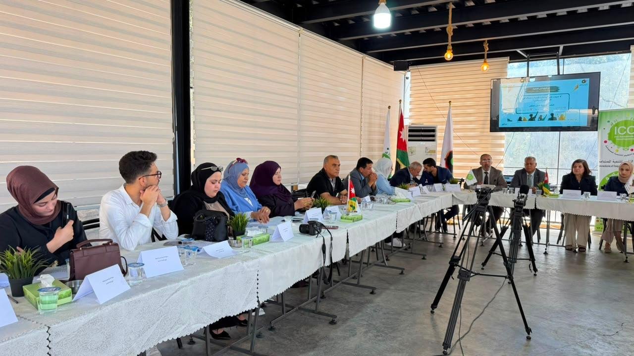 Panel-style meeting with many attendees seated along a long table in a bright, airy room; flags stand behind the speakers and a projector screen displays a presentation.