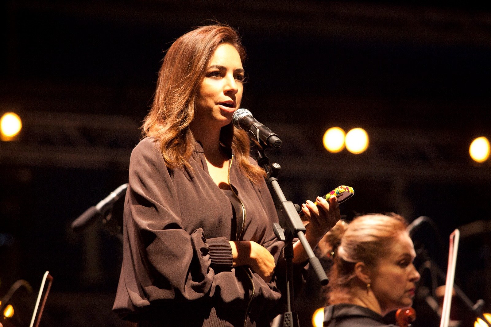 WhatsApp Image 2026-08-10 at 45552 PM 2 - عروبة الاخباري | Oroba News Female singer on stage speaking or singing into a microphone, wearing a brown jacket, stage lights in the background.
