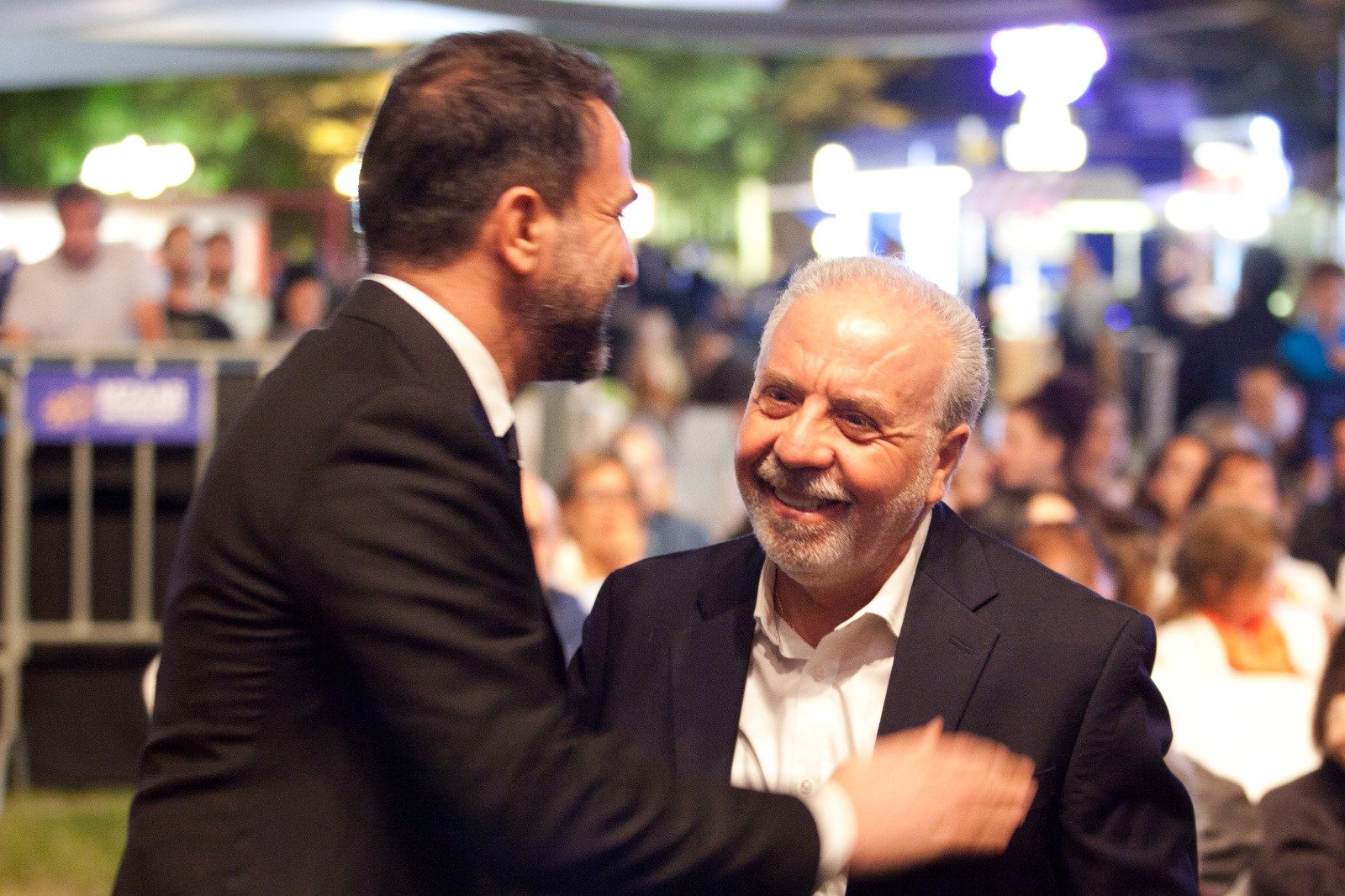 WhatsApp Image 2026-08-10 at 45552 PM 3 - عروبة الاخباري | Oroba News Two men in suits smiling and shaking hands at a crowded evening event in a party setting.