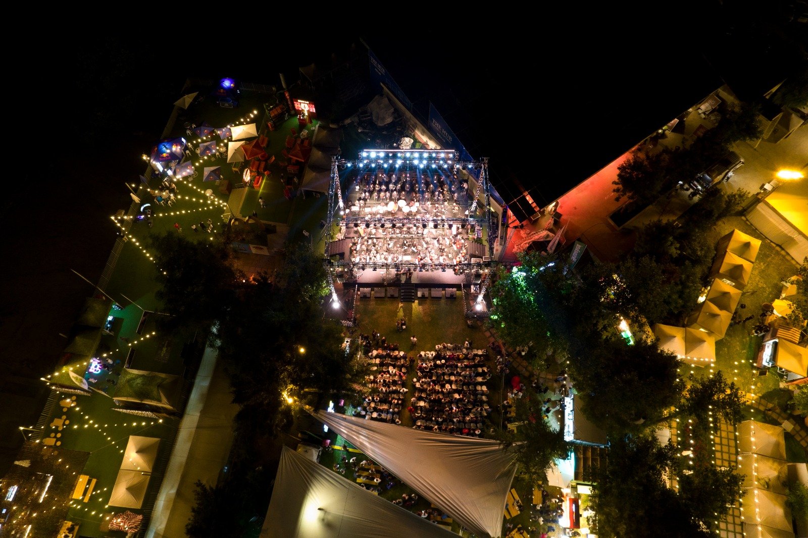 WhatsApp Image 2026-08-10 at 45552 PM 4 - عروبة الاخباري | Oroba News Aerial night view of an outdoor festival with a lit concert stage, surrounding tents, and a crowd watching the performance. - Oriented central stage with bright lights and audience seated on grass.