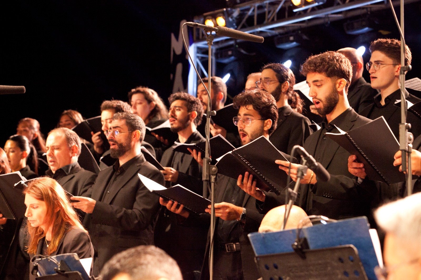 WhatsApp Image 2026-08-10 at 45554 PM 1 - عروبة الاخباري | Oroba News A choir of men and women in black outfits singing from sheet music on a stage with microphones and stage lights above them.