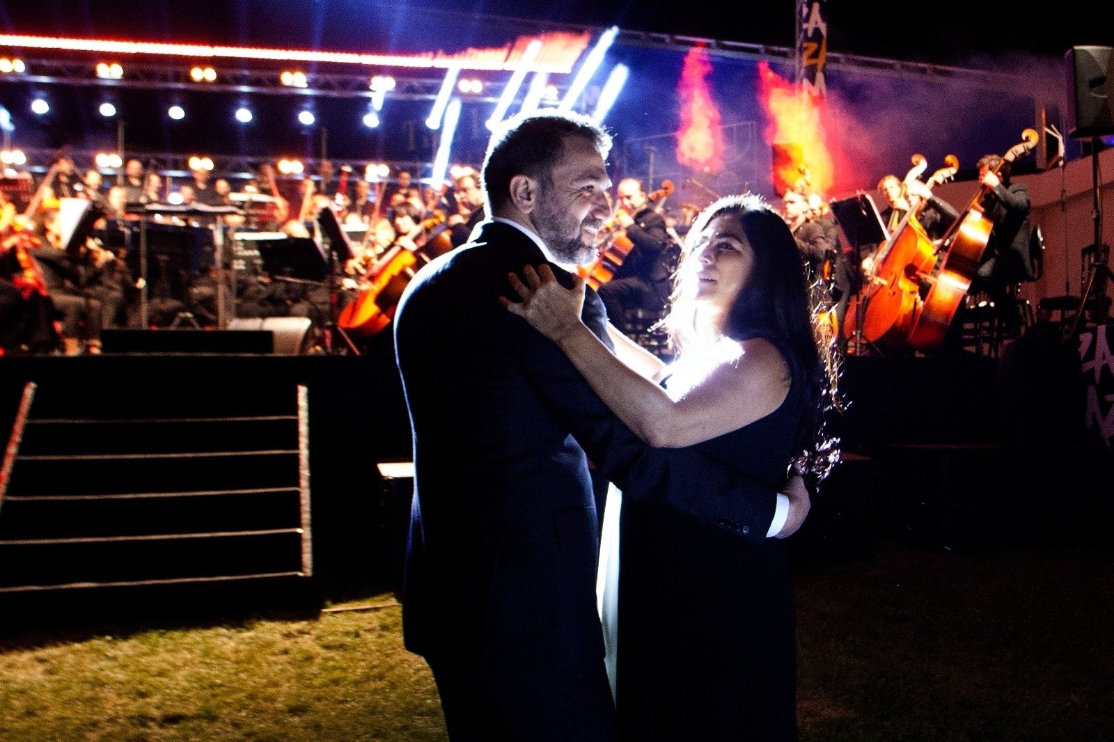 WhatsApp Image 2026-08-10 at 45554 PM 2 - عروبة الاخباري | Oroba News A couple dancing close together on a dark stage with an orchestra in the background under colorful lights, the man in a suit and the woman in a black dress smiling at each other.