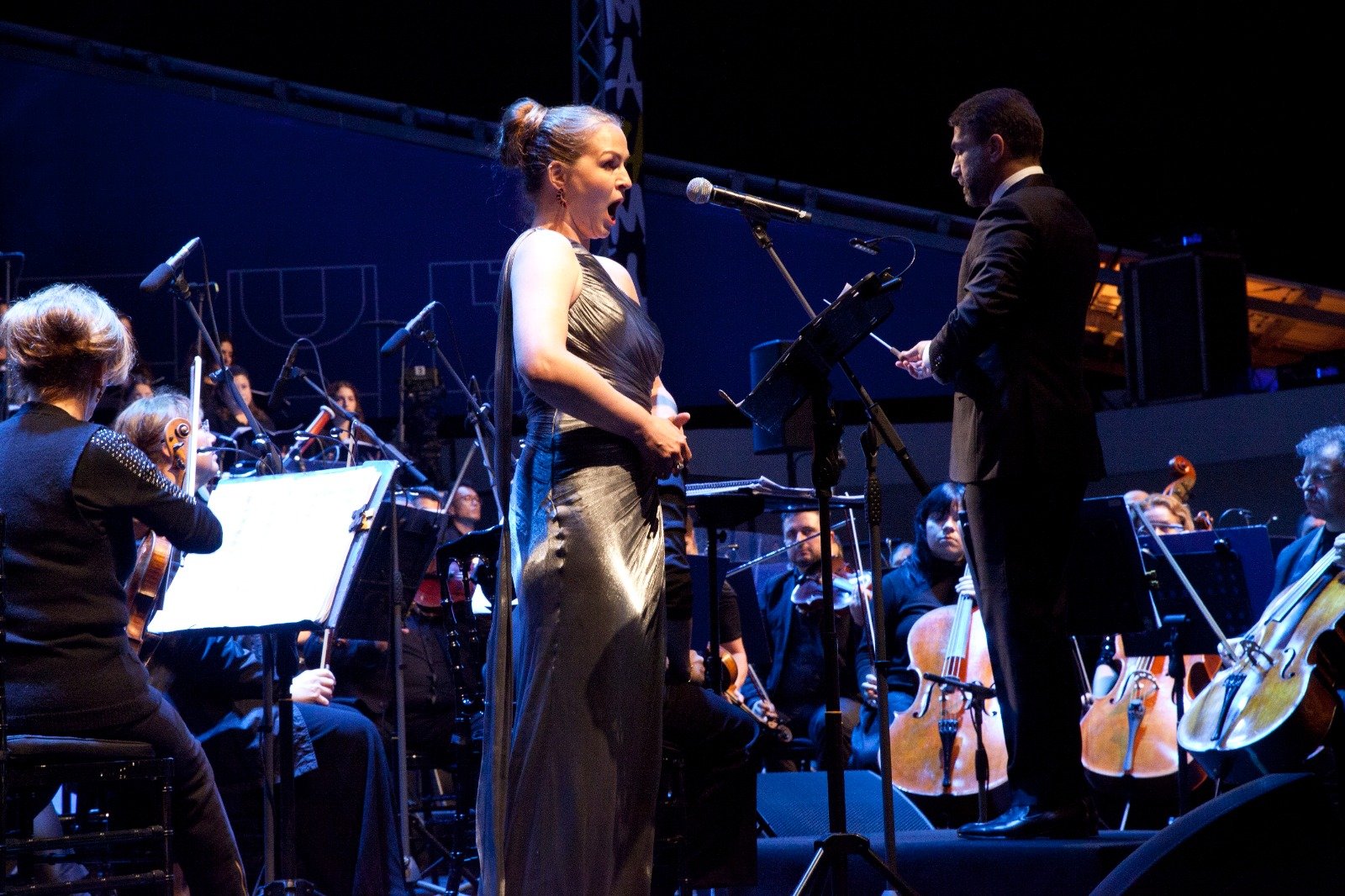 WhatsApp Image 2026-08-10 at 45554 PM 4 - عروبة الاخباري | Oroba News Female singer in a metallic gown performs with an orchestra on stage, mic in front of her, conductor nearby directing.