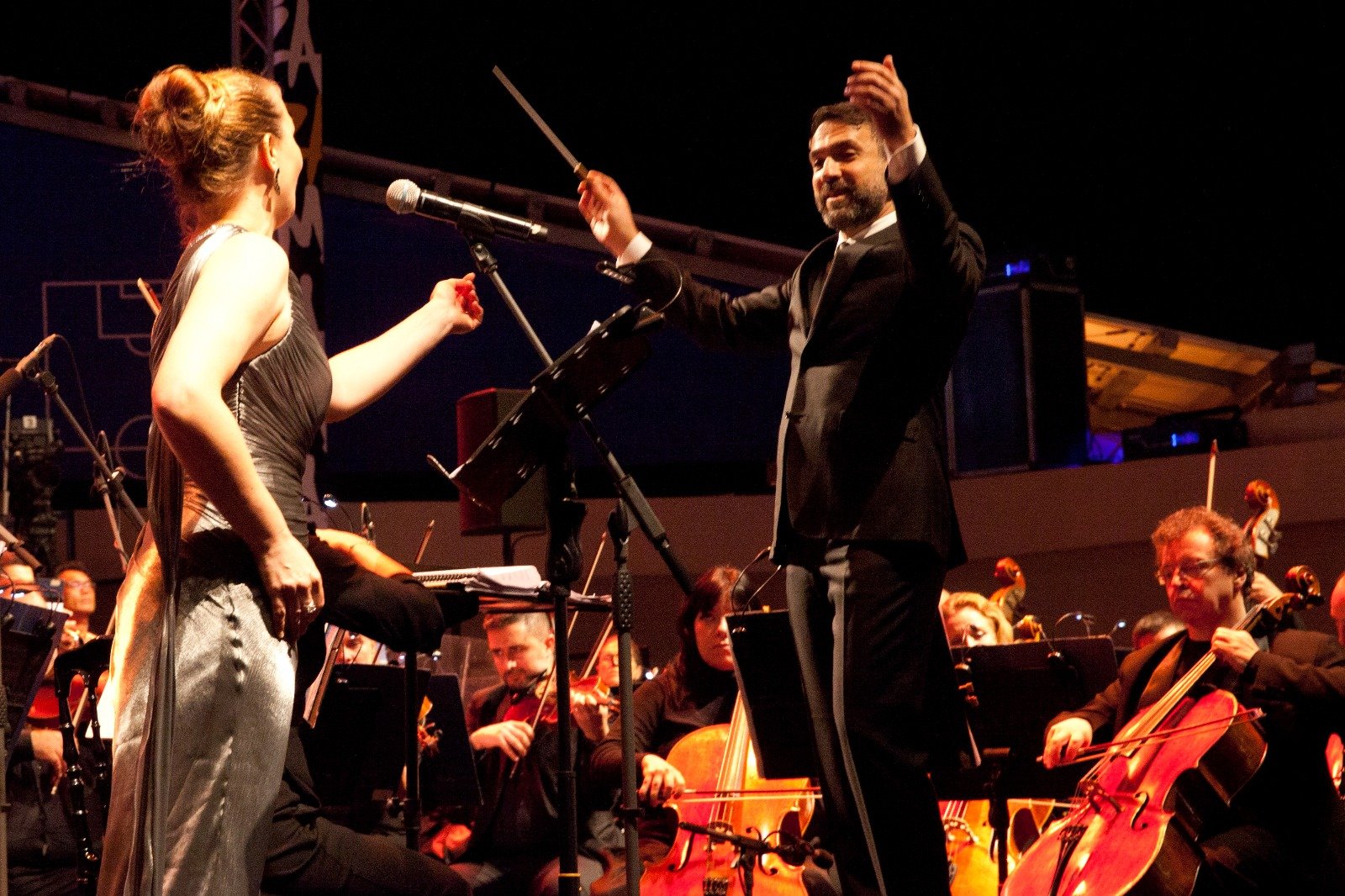 WhatsApp Image 2026-08-10 at 45554 PM 6 - عروبة الاخباري | Oroba News Conductor in a black suit raises his baton while a female singer in a silver dress performs at a microphone, with musicians and cellos behind them on stage.