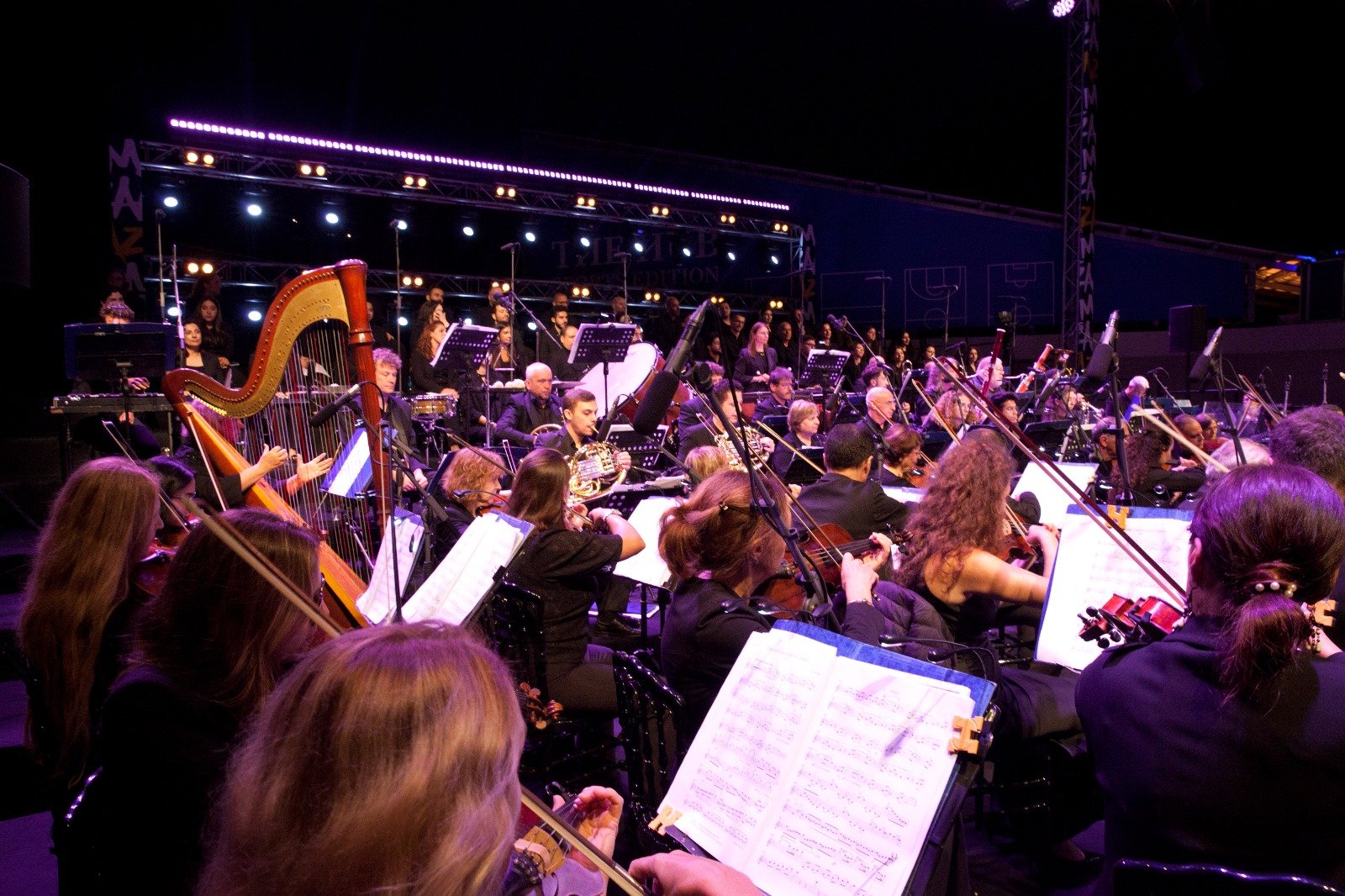 WhatsApp Image 2026-08-10 at 45554 PM 7 - عروبة الاخباري | Oroba News Full orchestra performing on a dark stage under purple lights, musicians playing strings, woodwinds, brass, and percussion with sheet music in front of them.