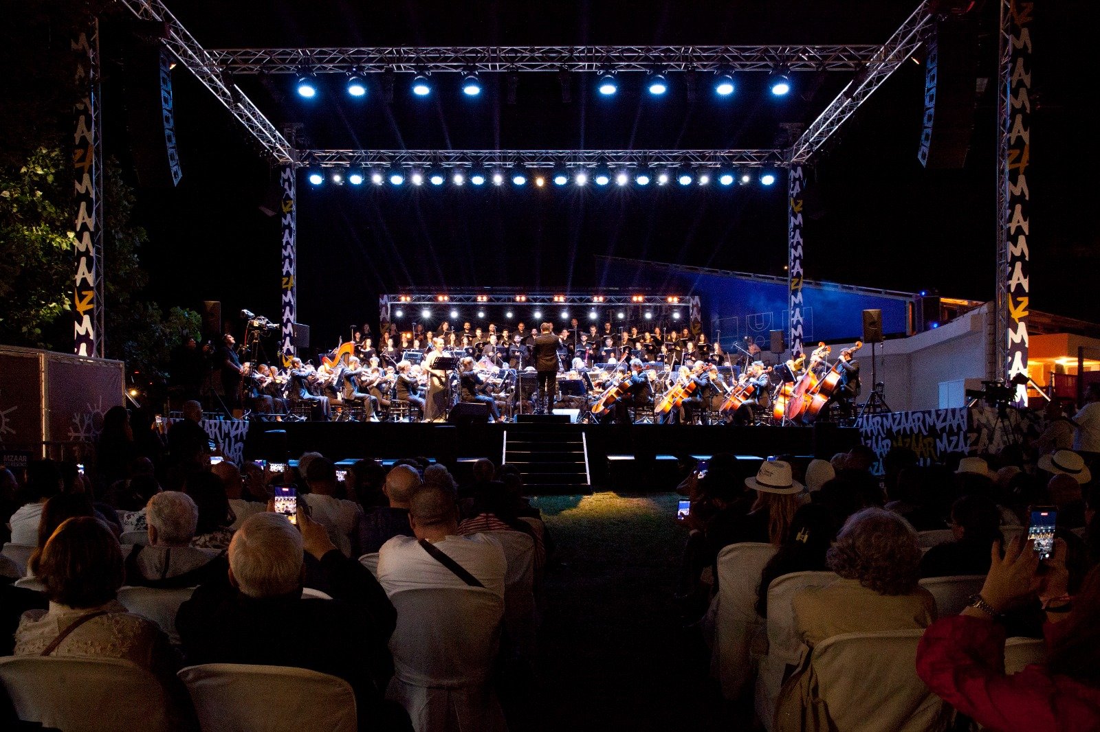 WhatsApp Image 2026-08-10 at 45554 PM - عروبة الاخباري | Oroba News Nighttime outdoor orchestra concert with a conductor leading string players on a large illuminated stage.