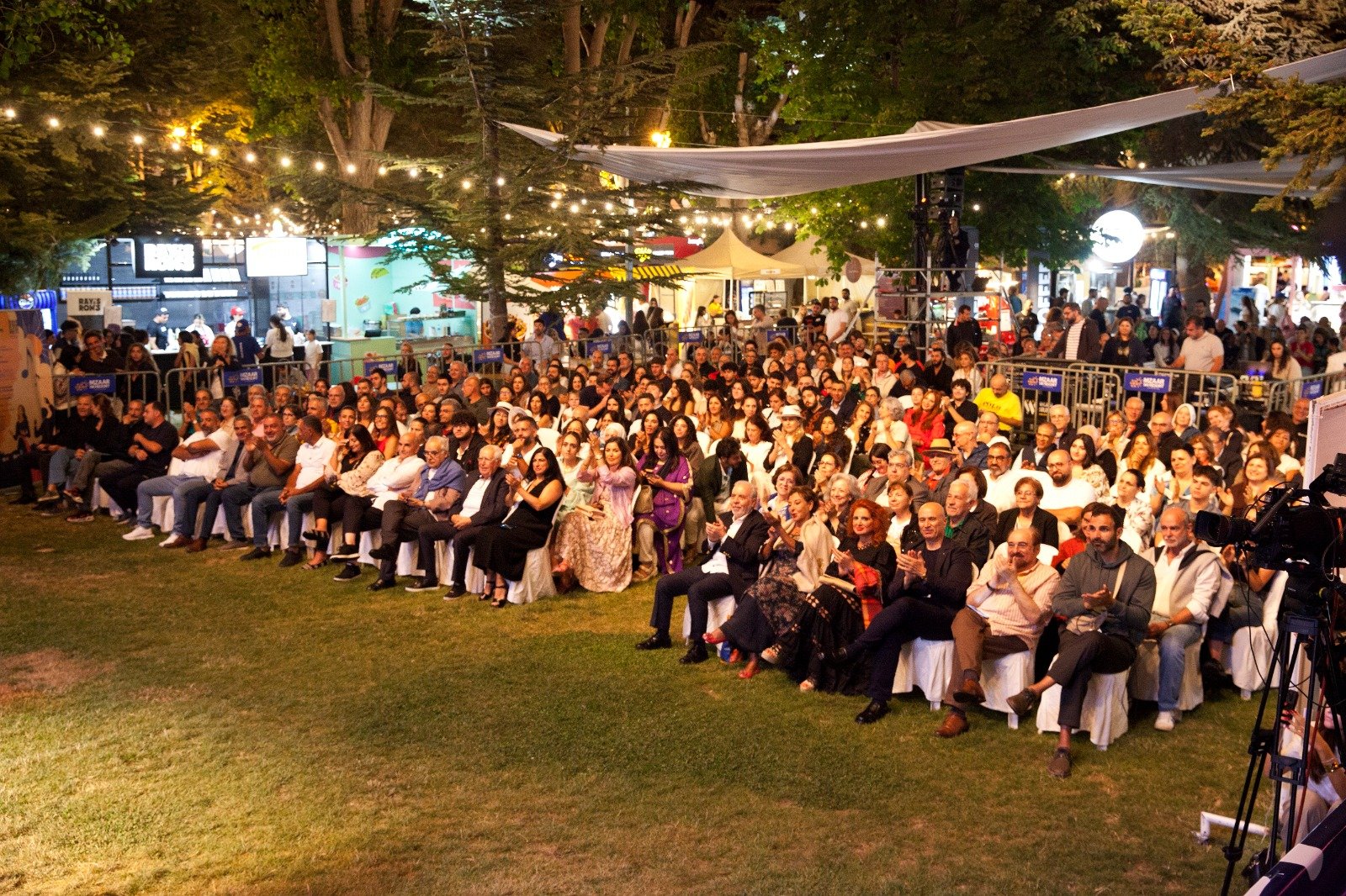 WhatsApp Image 2026-08-10 at 45555 PM 3 - عروبة الاخباري | Oroba News Crowd of people seated in rows on a grassy outdoor event at night, string lights overhead and festival stalls behind.