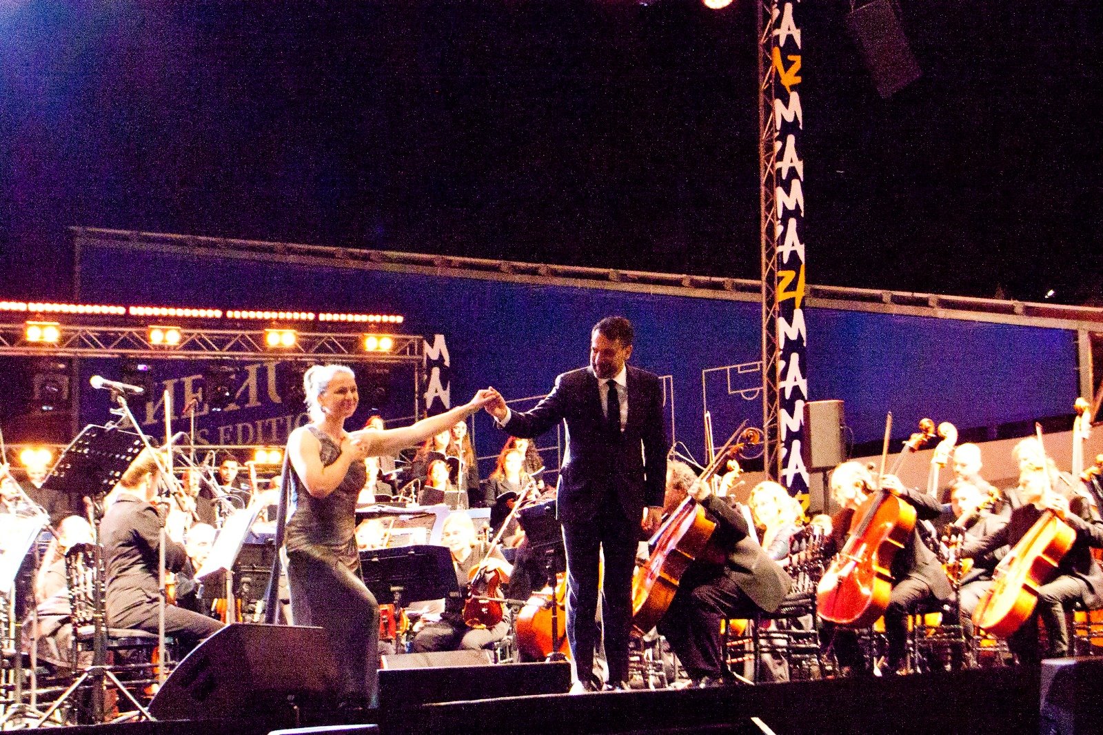 WhatsApp Image 2026-08-10 at 45555 PM 5 - عروبة الاخباري | Oroba News Conductor in a suit and a female soloist shake hands on a concert stage as a full orchestra plays behind them.