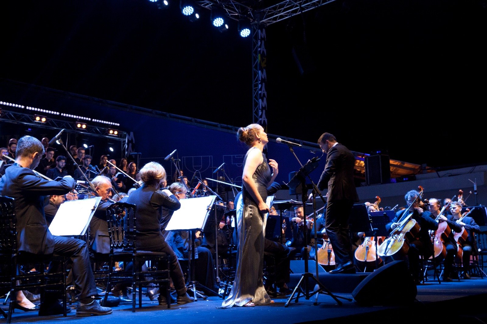 WhatsApp Image 2026-08-10 at 45555 PM - عروبة الاخباري | Oroba News Female soloist in a silver gown sings into a microphone while an orchestra accompanies on stage during a concert, blue lighting overhead.