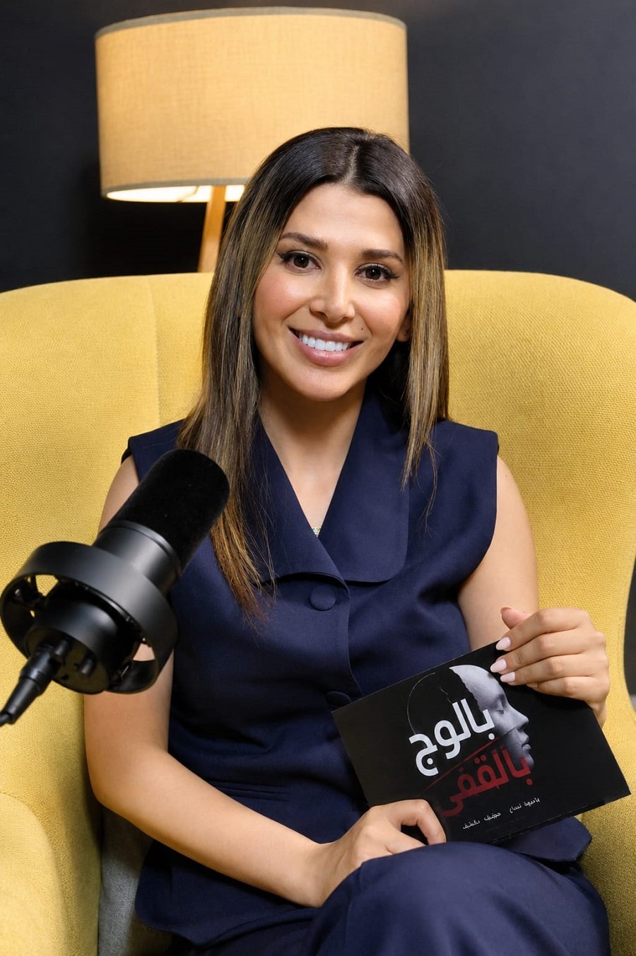 Smiling woman in a navy dress sits in a mustard-yellow chair, speaking into a large microphone and holding a black booklet with Arabic text in her lap.
