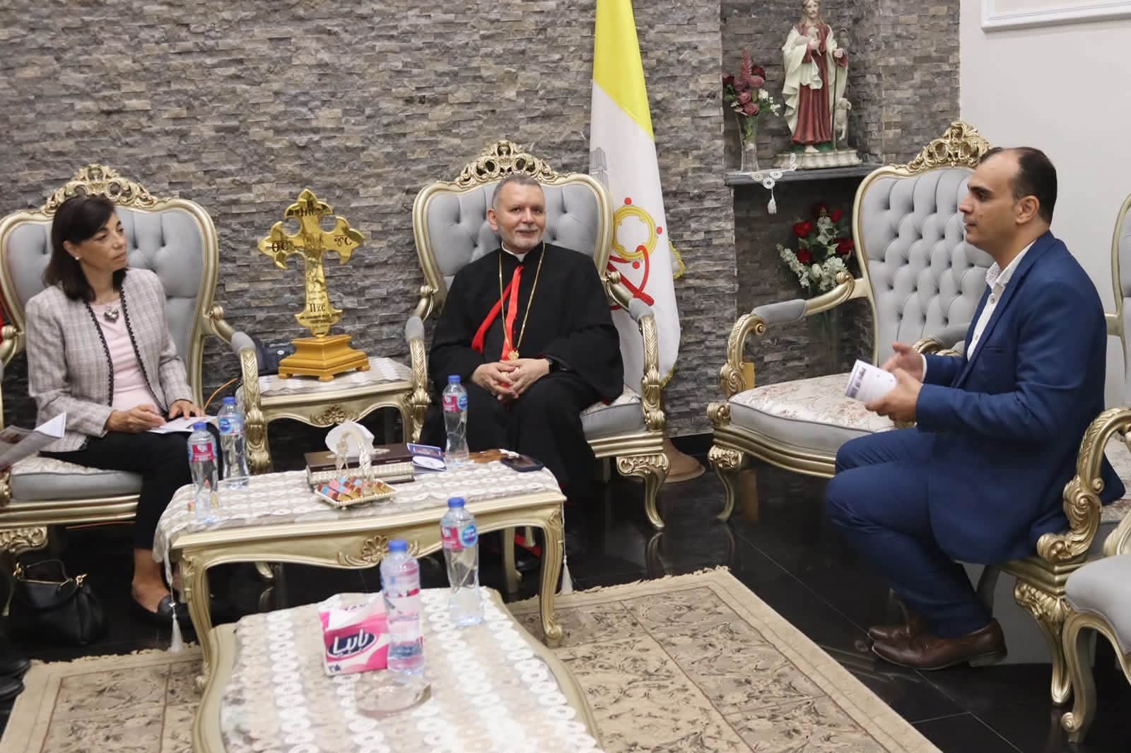 Three people sit in gold-trimmed chairs around a coffee table during a formal meeting; a clergyman in black robes with a red sash sits center.