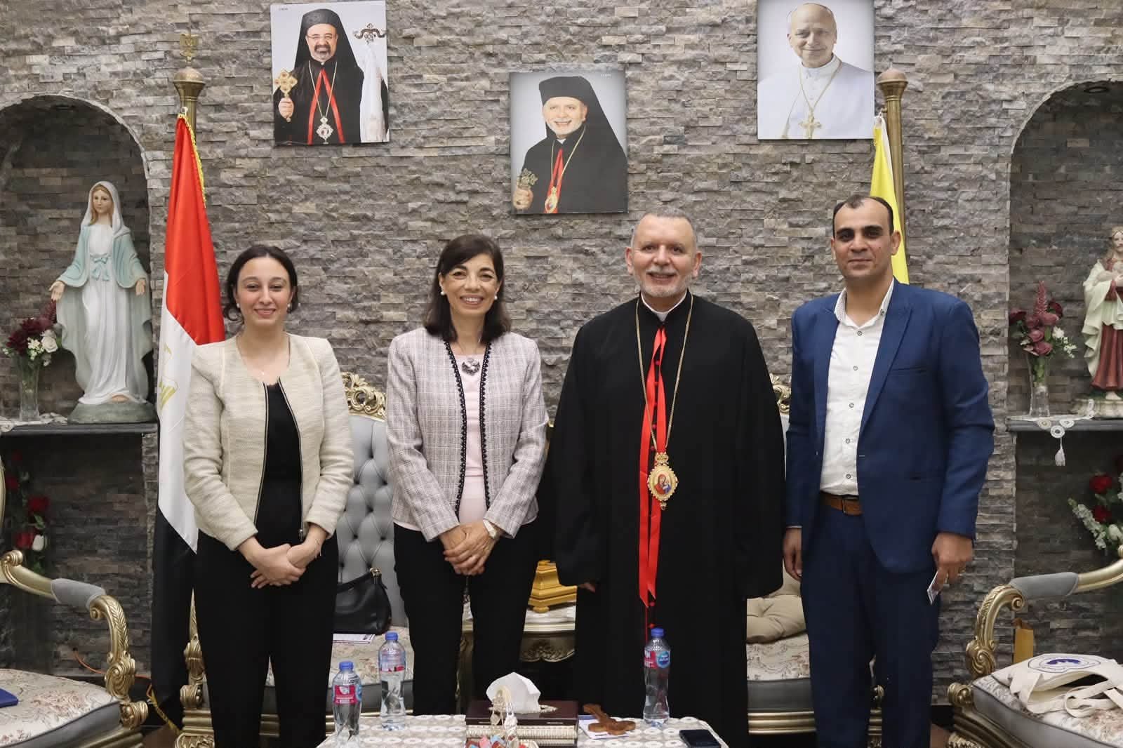 Four people pose for a formal photo in a religious setting: two women, a priest in black robes with red trim, and a man in a blue suit, standing in front of a stone wall with religious portraits and statues.