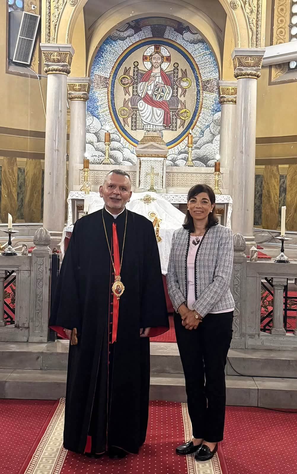 Priest in black robes with red sash and medallion posing beside a woman inside a church with ornate mosaic behind them.
