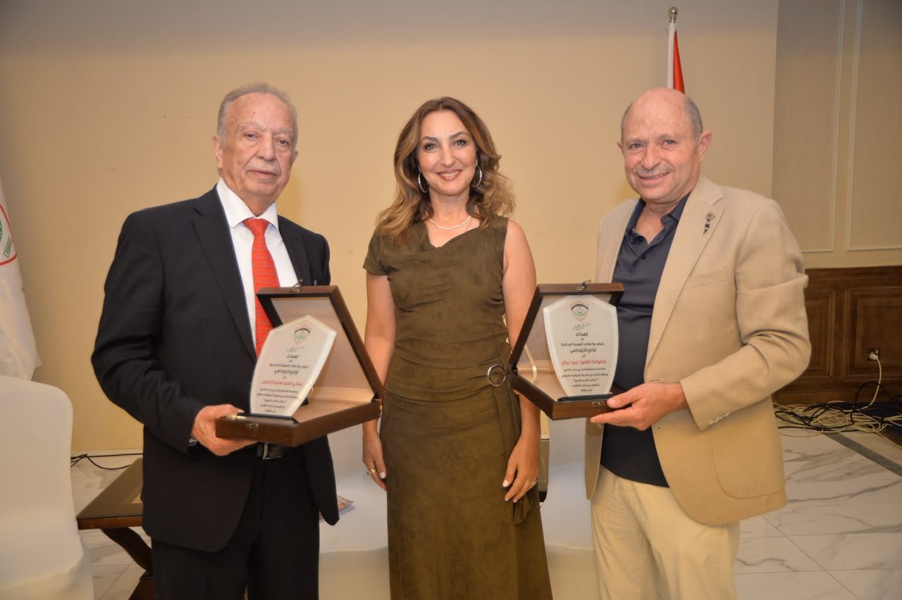 Two men in suits hold award plaques while a smiling woman stands between them at an indoor awards ceremony.