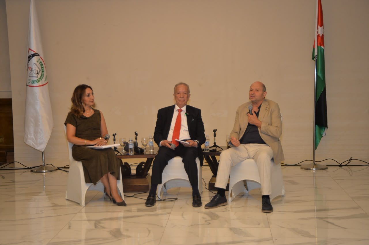 Panel discussion with three speakers seated on a stage, flags on either side, glassware and microphones on the table in front of them.