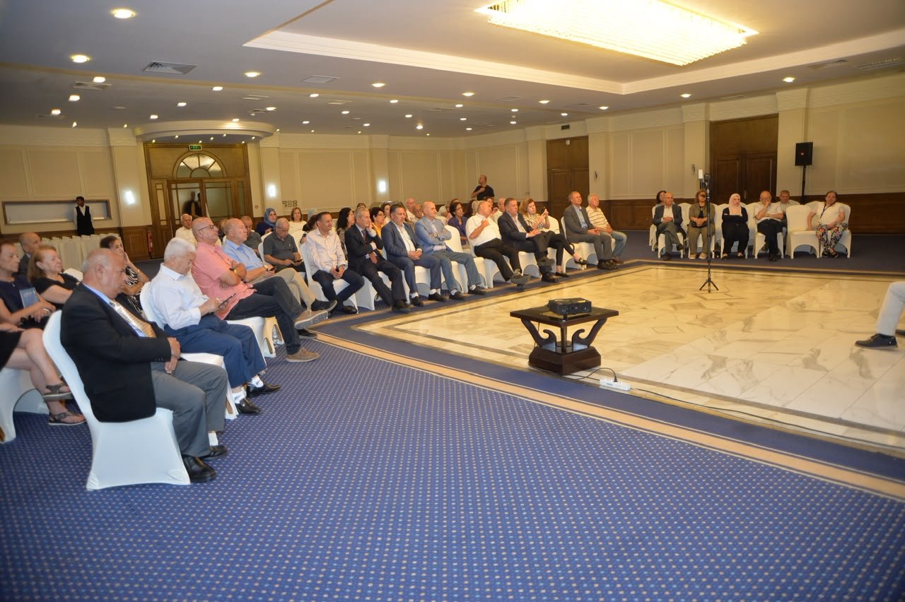 Audience seated in rows in a formal event hall, watching a presentation with a small projector on a table at the front and a speaker area on a marble-floored stage.