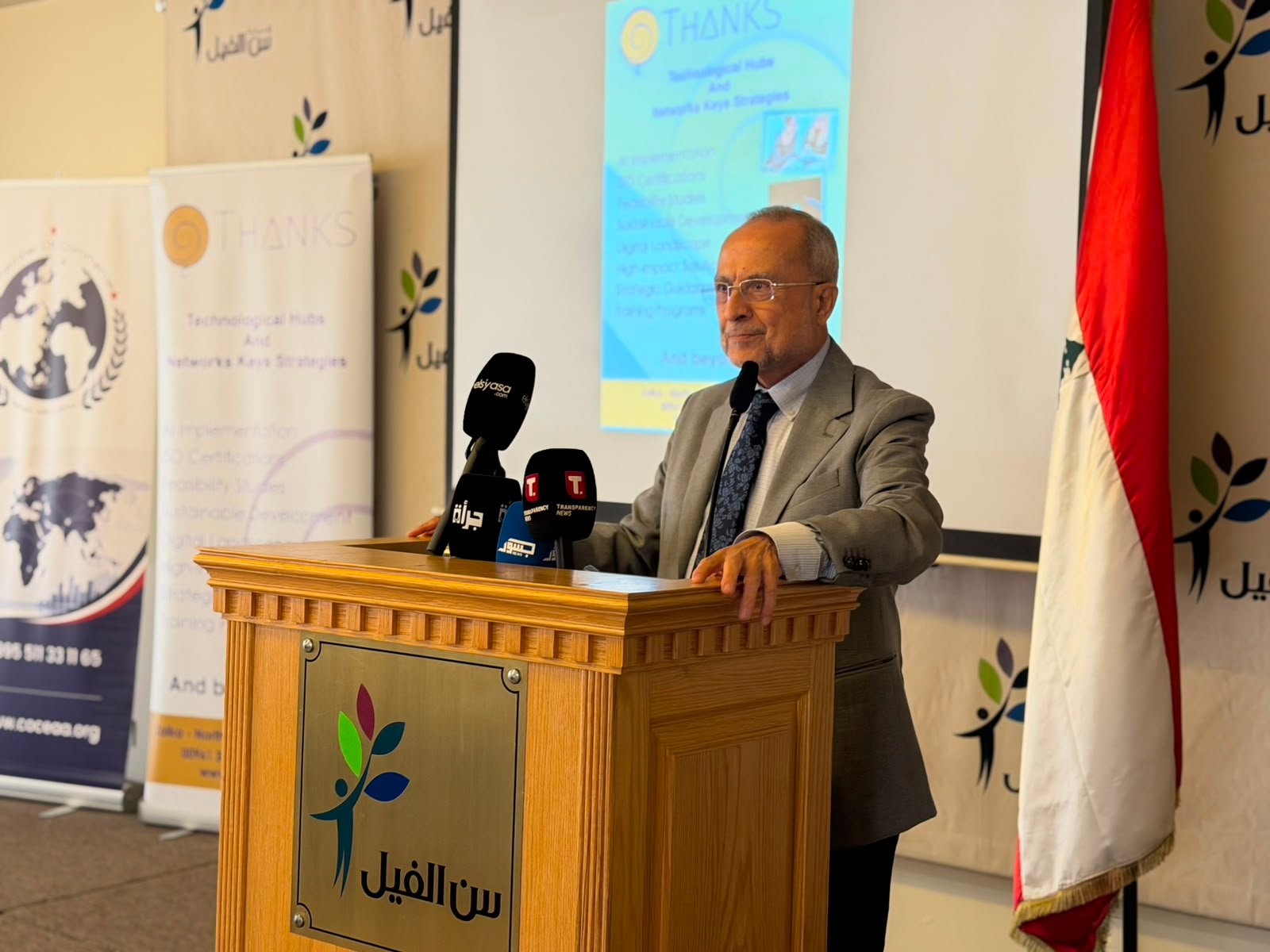 A man in a gray suit giving a speech at a wooden podium with several microphones, banners, and an Iraqi flag in the background.
