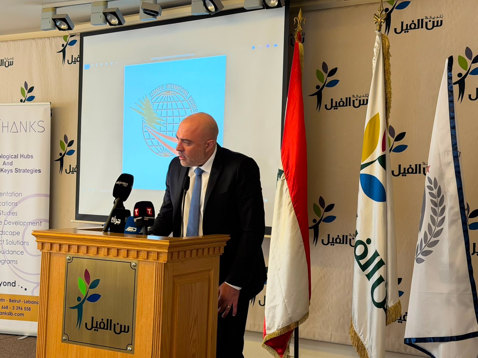 A man in a suit giving a speech at a wooden podium with multiple microphones, standing in front of a projection screen and national/organizational flags.