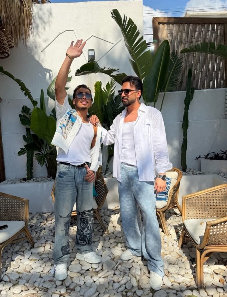 WhatsApp Image 2026-08-13 at 34444 PM - عروبة الاخباري | Oroba News Two men in white shirts and jeans pose outdoors among tropical plants; one raises his hand in the air.