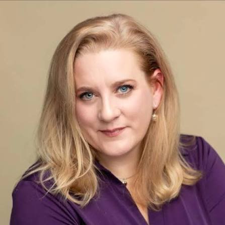 Head-and-shoulders portrait of a woman with blonde hair and blue eyes, wearing a purple blouse, looking at the camera with a gentle smile.