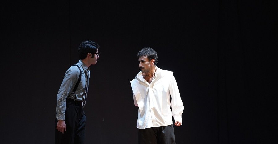 Two men on a stage face off in a tense confrontation; one wears suspenders and glasses, the other a loose white shirt on a dark backdrop.