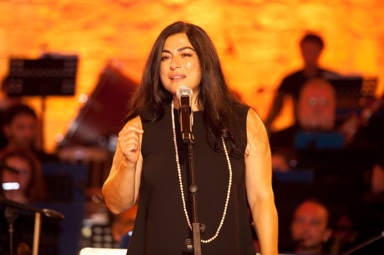 Female singer in a sleeveless black dress with a pearl necklace performs into a microphone on a warmly lit stage.