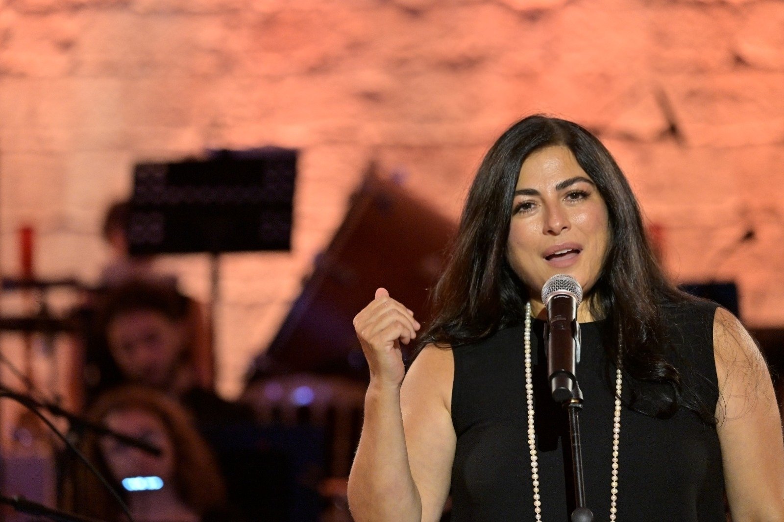 Singer on stage in a black dress with a pearl necklace, singing into a microphone against a warm brick backdrop.
