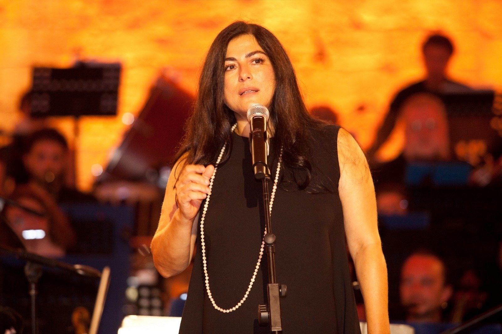 A female singer in a black dress with a long pearl necklace, performing into a microphone on a warm orange-lit stage.