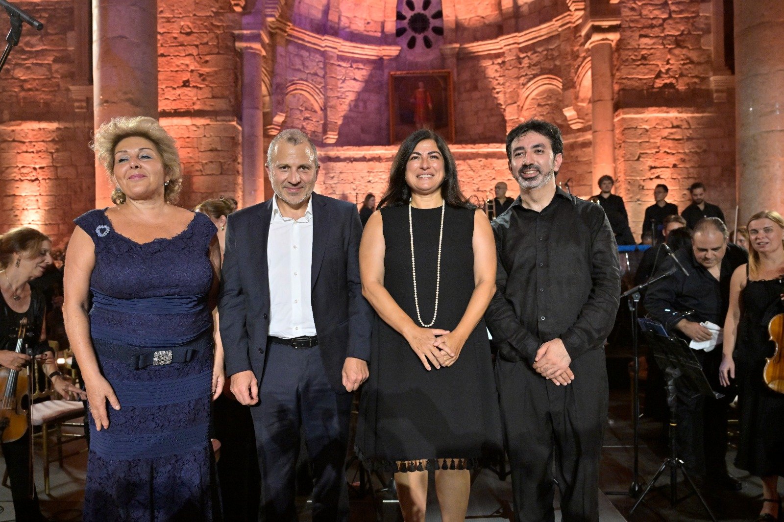 Four people in formal attire posing on a stage with an orchestra in the background.