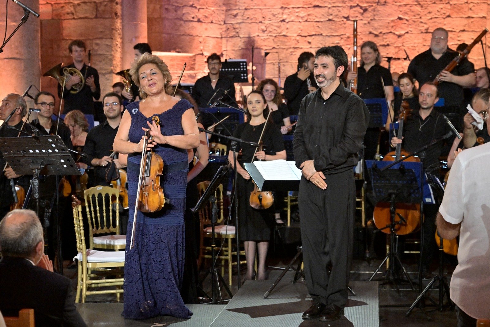 Violinist in a blue lace gown stands with her violin and bow at the front of a stage, a male musician beside her, as a full orchestra and audience watch behind them.