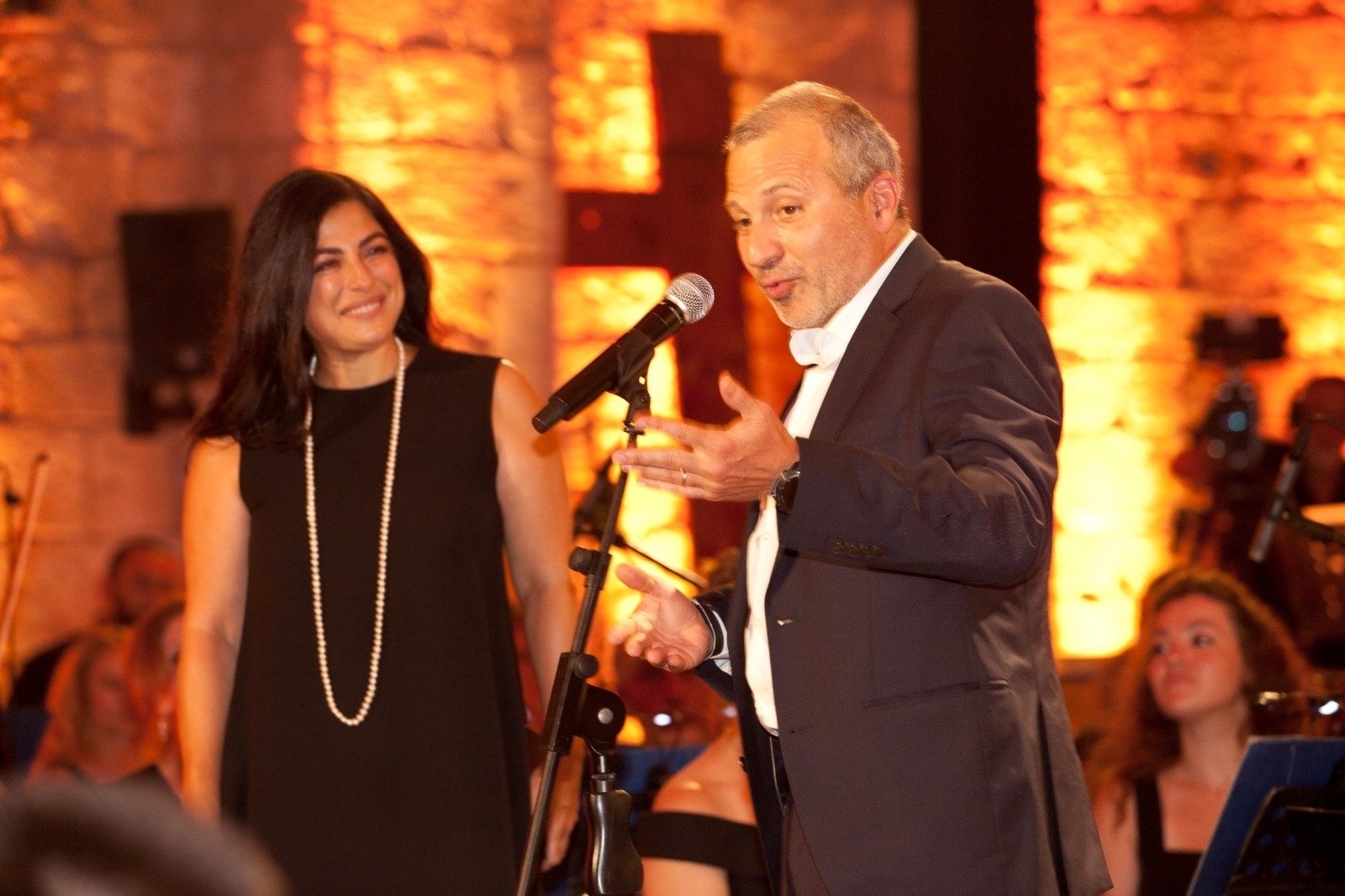 Man in tuxedo singing into microphone on stage with a smiling woman in black nearby; warm orange stage lights in background.