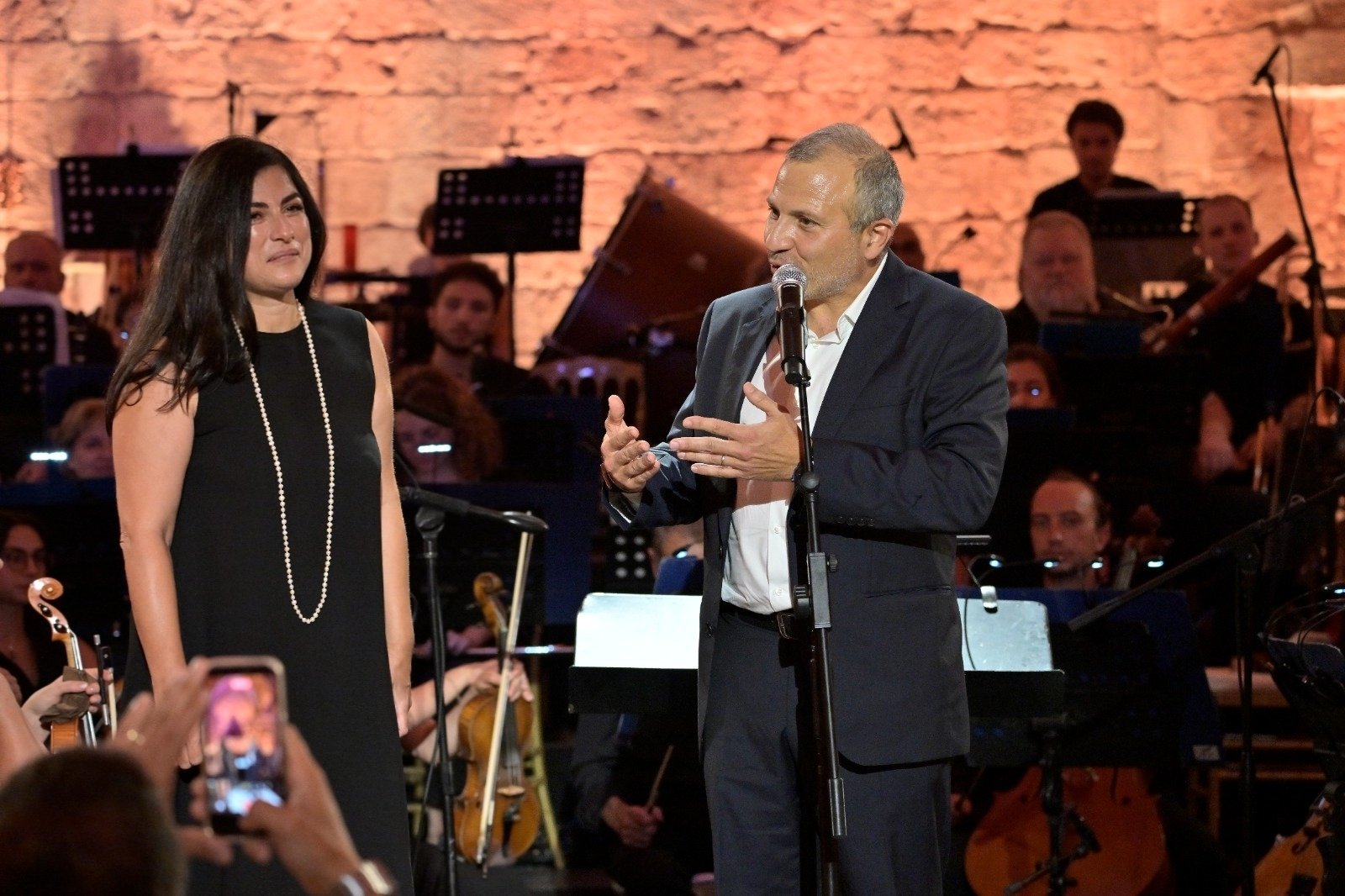 Man in a dark suit speaks into a microphone on stage beside a woman in a black dress with a pearl necklace, with an orchestra behind them.