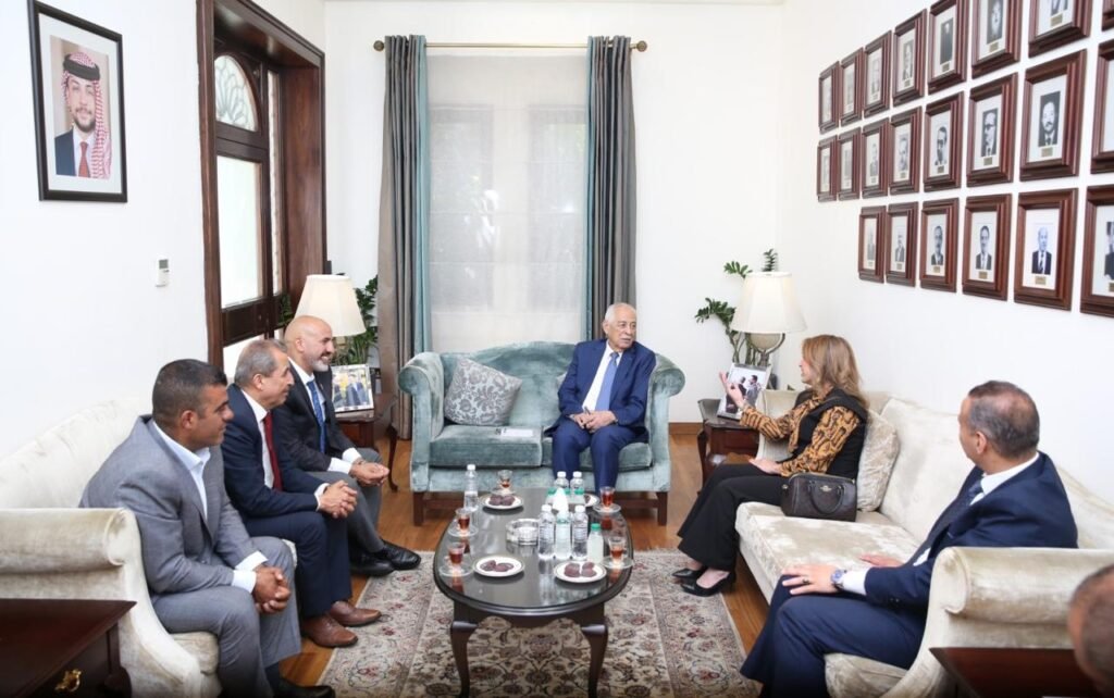 WhatsApp Image 2026-08-19 at 25221 PM - عروبة الاخباري | Oroba News Formal meeting in a living room: six people seated around a glass coffee table, central man in a blue suit on a blue sofa, wall lined with framed portraits.