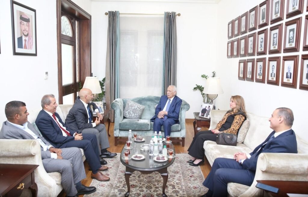 WhatsApp Image 2026-08-19 at 25221 PM1 - عروبة الاخباري | Oroba News Six professionals in formal attire sit around a coffee table in a bright living room, with an older man in a blue suit centered on a teal sofa; framed photos line the wall. - Informative scene context for a meeting image