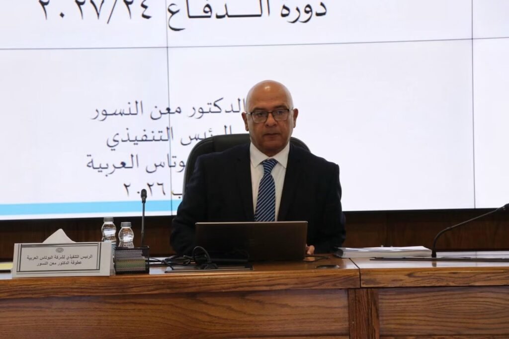 WhatsApp Image 2026-08-19 at 52103 PM1 - عروبة الاخباري | Oroba News Bald man in a suit delivering a presentation at a conference, with a laptop and microphone on the desk and Arabic text on a large screen behind him.