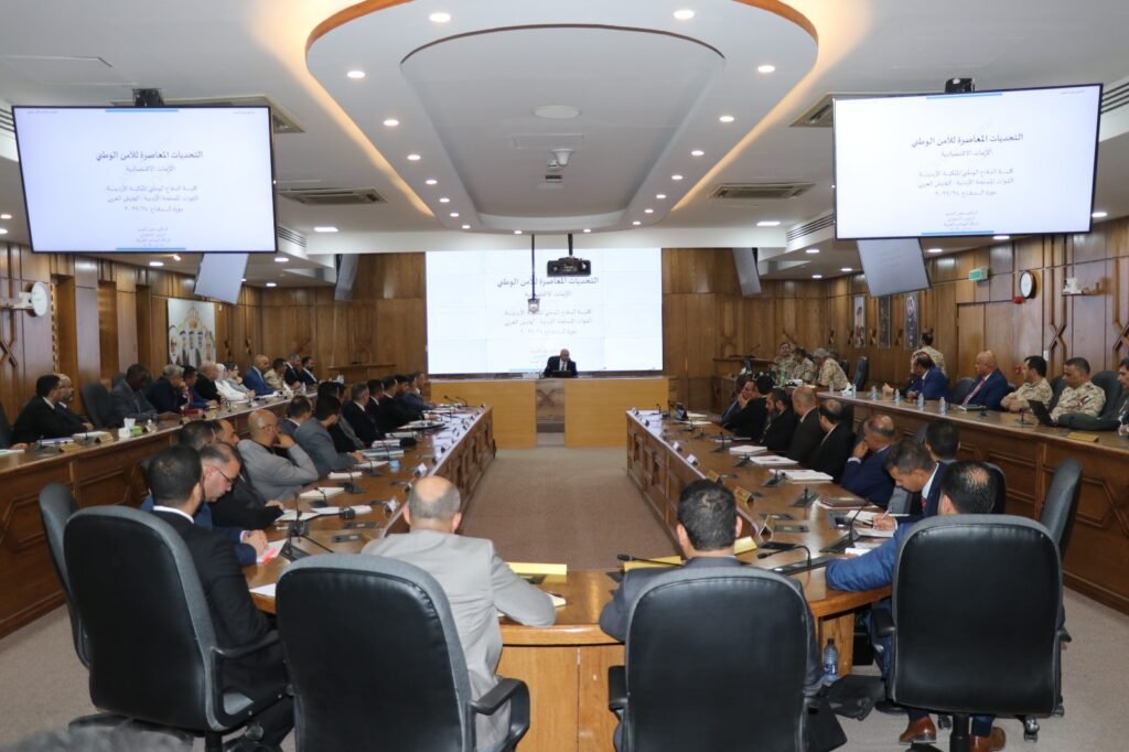 WhatsApp Image 2026-08-19 at 52103 PM2 - عروبة الاخباري | Oroba News Full view of a formal meeting in a large conference room with attendees around a U-shaped wooden table; a speaker sits at the front beneath a large screen displaying a presentation.