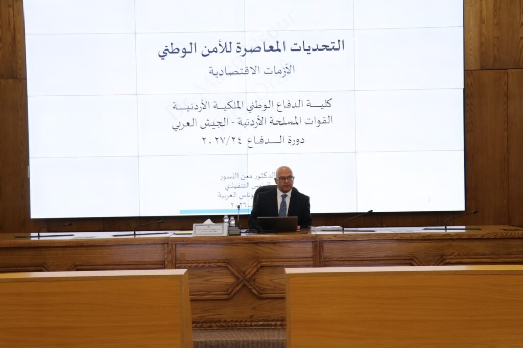 WhatsApp Image 2026-08-19 at 52103 PM3 - عروبة الاخباري | Oroba News Man in a suit seated at a podium in a conference room, with a large white Arabic presentation on screen behind him.