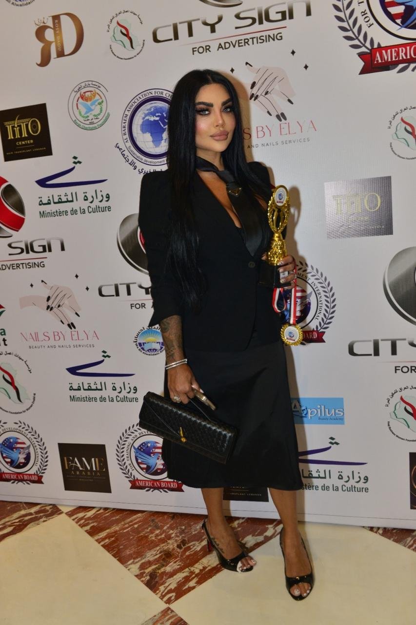 WhatsApp Image 2026-08-20 at 15825 AM2 - عروبة الاخباري | Oroba News Woman with long black hair in a black outfit posing with a gold trophy on a backdrop of logos; holding a black clutch and wearing black heels.