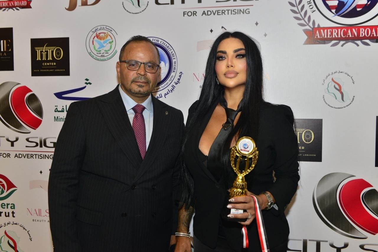 WhatsApp Image 2026-08-20 at 15826 AM - عروبة الاخباري | Oroba News Man in a dark suit and a woman in a black blazer pose with a gold trophy at an award event backdrop.
