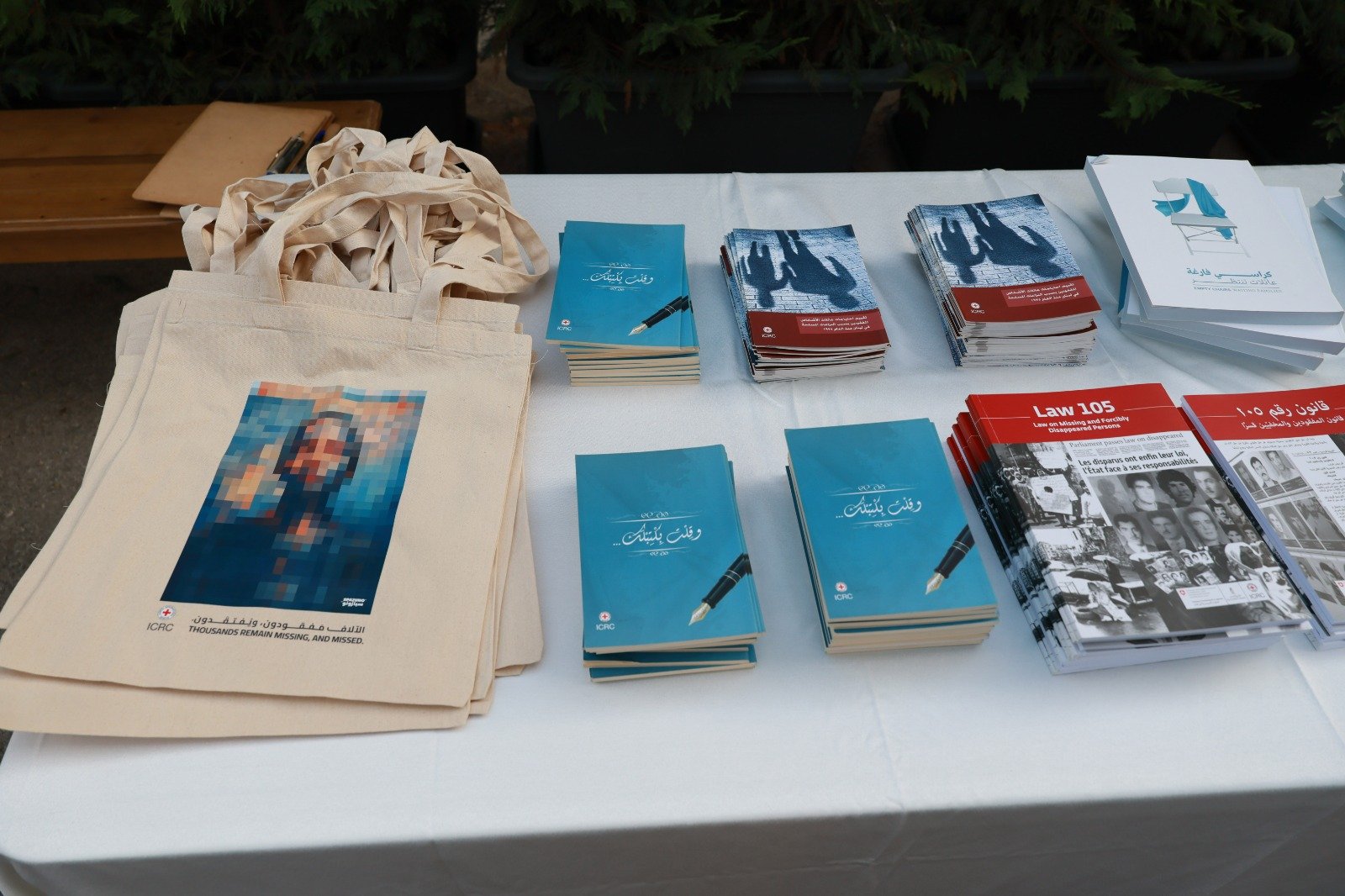 Beige tote bags with a pixelated portrait on the front, arranged on a table beside blue booklets and red pamphlets on a white tablecloth.