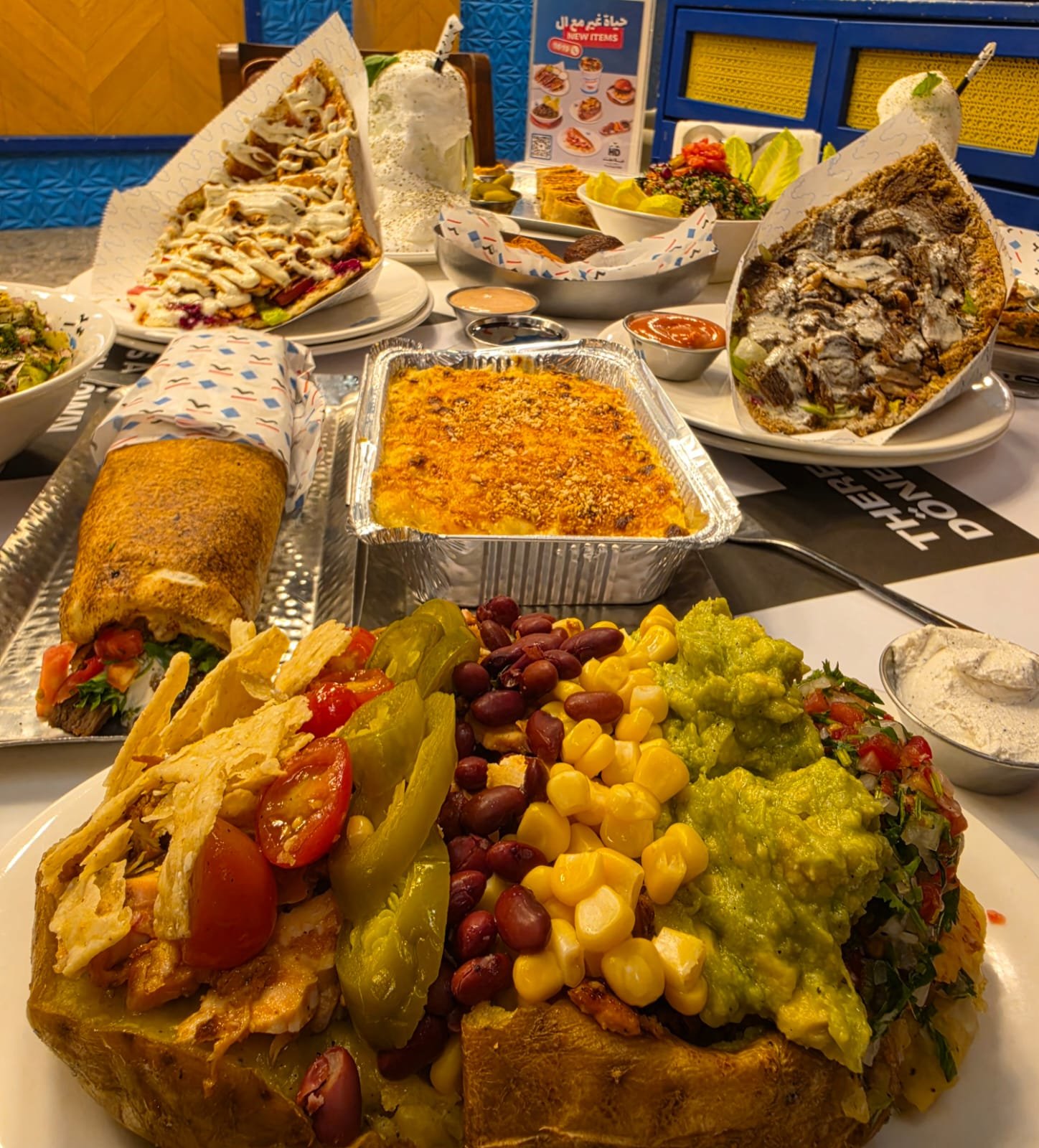Vibrant crowded buffet with stuffed bread bowls, a taco wrap, corn, beans, guacamole, and a cheesy baked dish on a table.