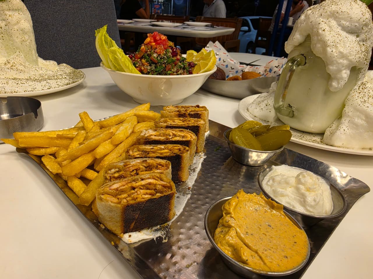 Platter with sliced wrap rolls, French fries, pickles, and three dipping sauces on a metal tray; background bowls and a foamy beverage.