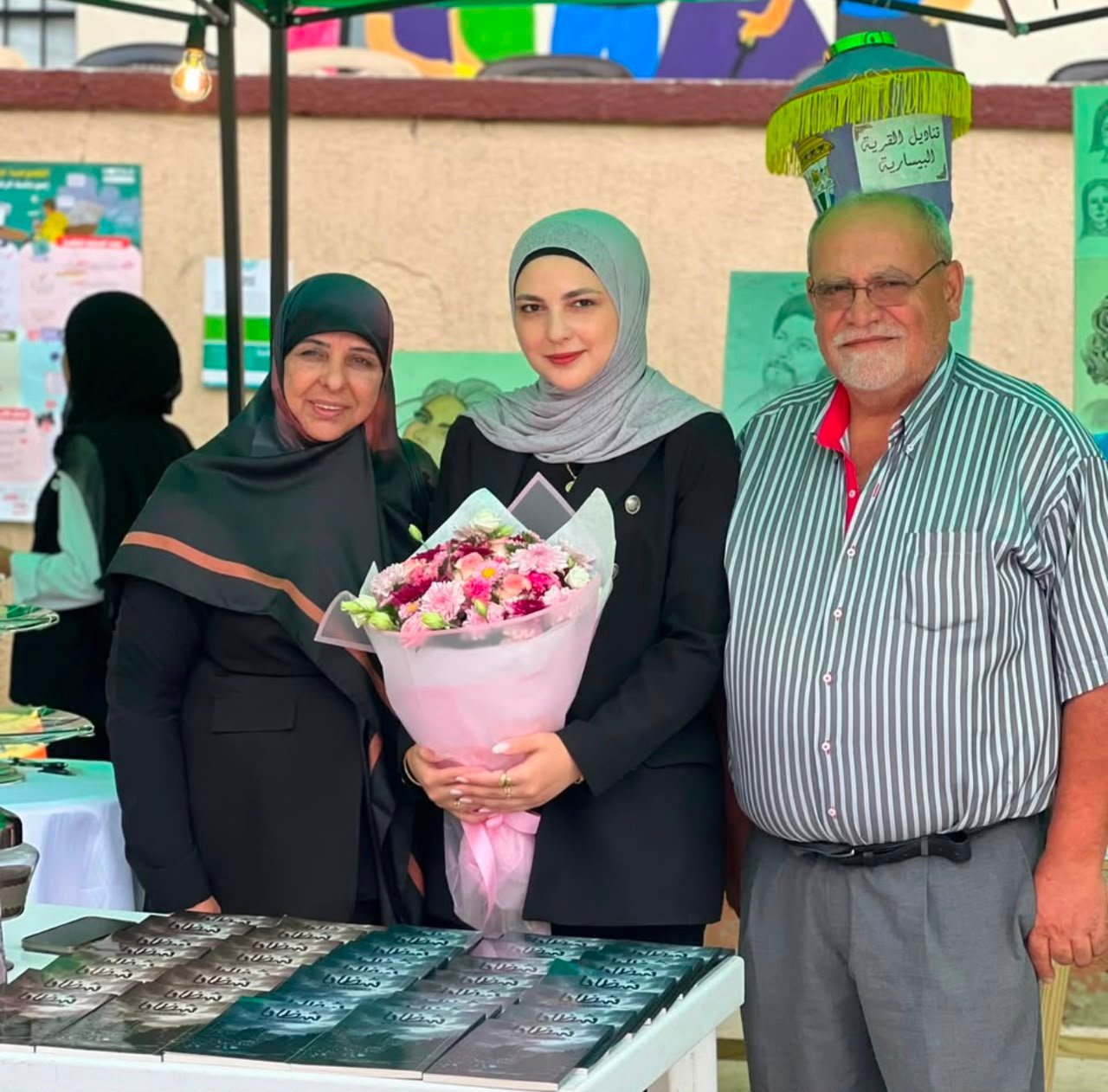 Center woman in a grey hijab holding a pink bouquet, flanked by a woman in a dark hijab and a man in a striped shirt at a booth with a table of books.