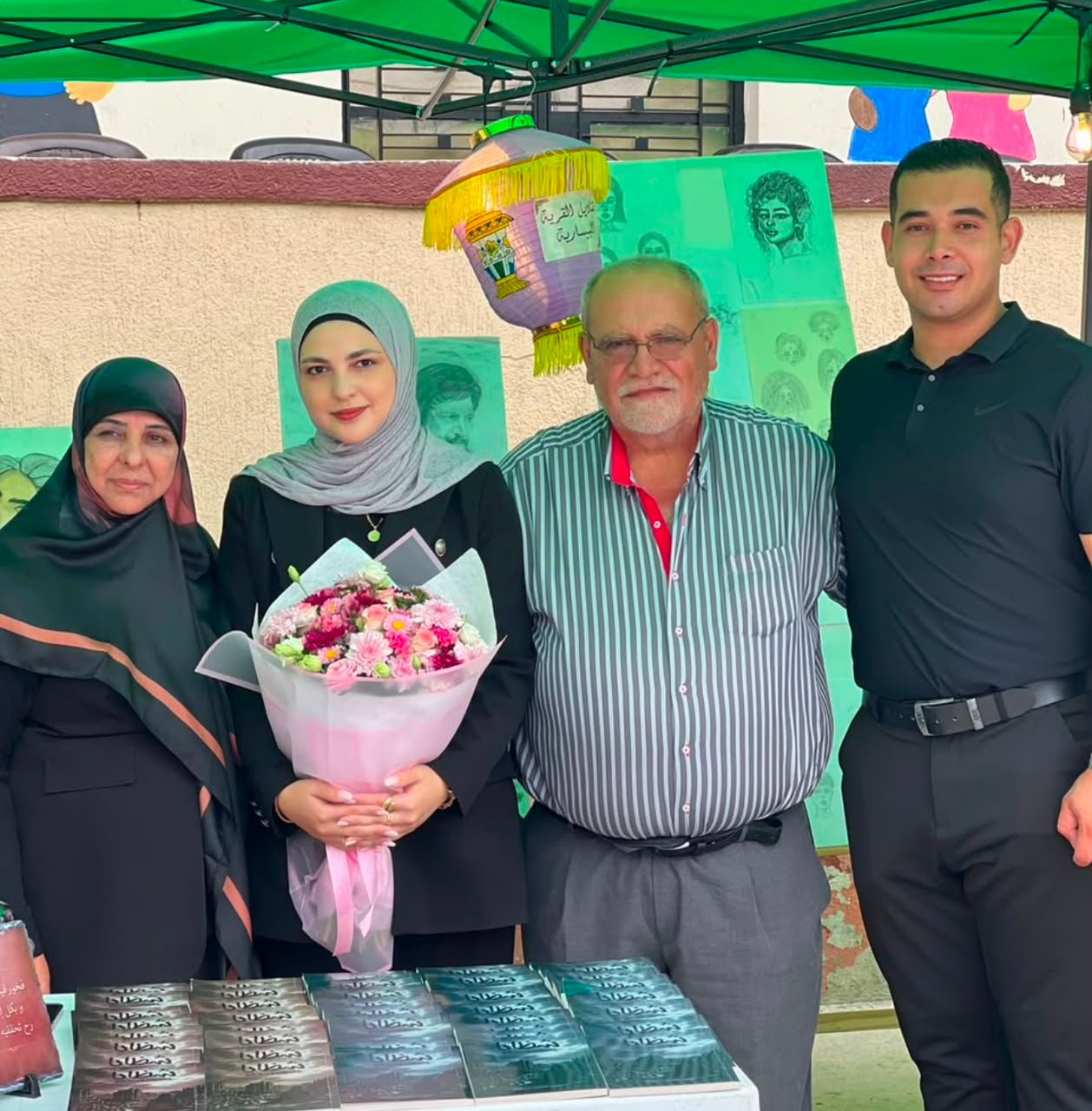 Four people under a green canopy at a book stall; the woman in a hijab holds a bouquet while books are laid out on a table in front.