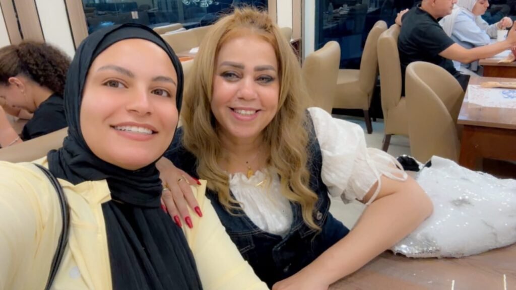 WhatsApp Image 2026-08-22 at 33226 PM1 - عروبة الاخباري | Oroba News Two women smile for a selfie in a restaurant; one wears a black hijab and pale yellow shirt, the other has blonde hair and a white blouse with a denim vest in front.